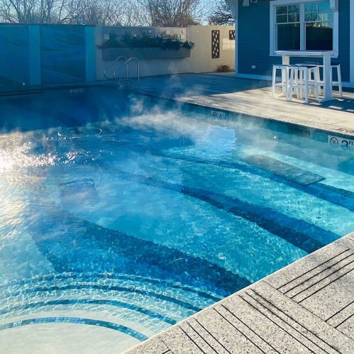 A serene swimming pool with steam rising, surrounded by a patio and greenery under a clear blue sky.