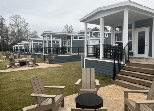 A row of modern cabins with porches, surrounded by green grass and wooden chairs in the foreground.