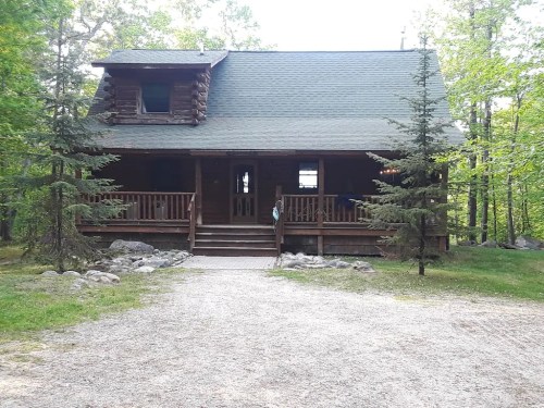 A rustic log cabin surrounded by trees, featuring a front porch and gravel pathway.