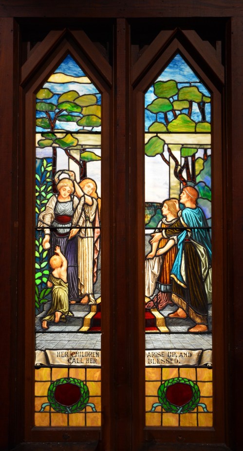 Stained glass window depicting a scene with women and children, surrounded by trees and vibrant colors.