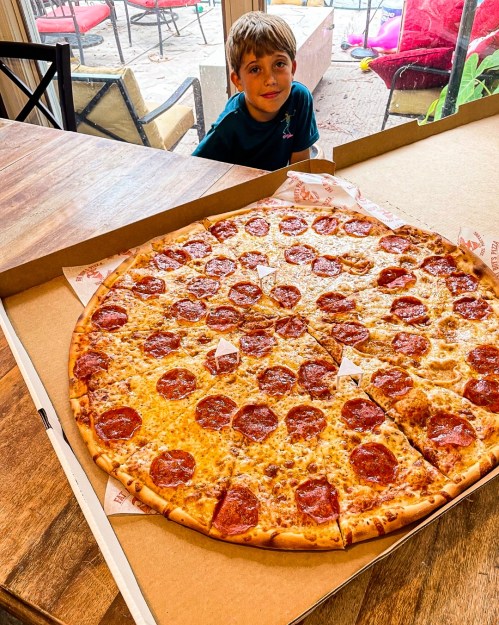 A large pepperoni pizza sits in a box, with a young boy peeking from behind it, looking excited.