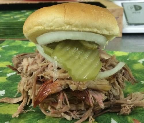 A pulled pork sandwich topped with pickles and onions, served on a soft bun, resting on a green leaf-patterned surface.