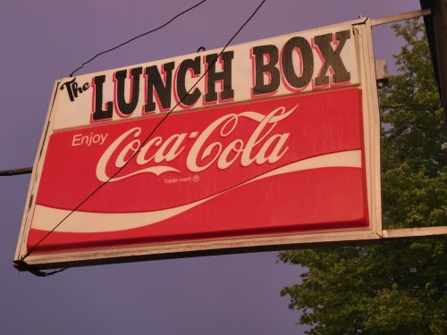 Sign for "The Lunch Box" featuring a Coca-Cola logo, set against a purple sky.