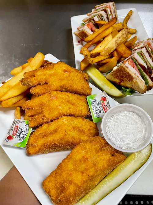 Two plates of food: one with golden fried fish, fries, and pickles; the other with a club sandwich and more fries.