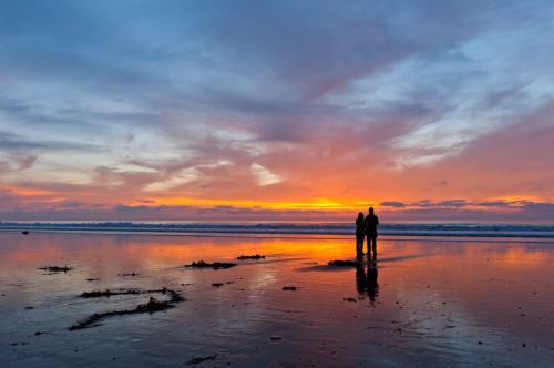 A couple stands on a beach at sunset, reflecting on the wet sand with vibrant colors in the sky.