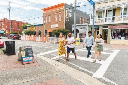 Four people walk across a crosswalk in a vibrant downtown area with shops and colorful buildings.