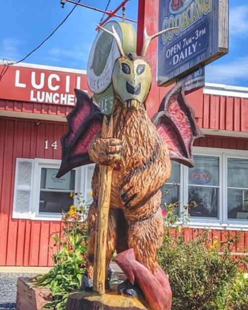 A wooden statue of a creature with wings and horns stands outside a restaurant, surrounded by greenery and a sign.