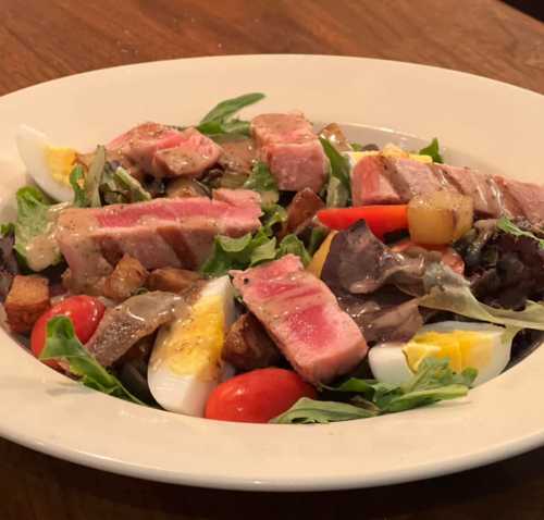 A colorful salad with mixed greens, sliced steak, cherry tomatoes, hard-boiled eggs, and croutons in a white bowl.