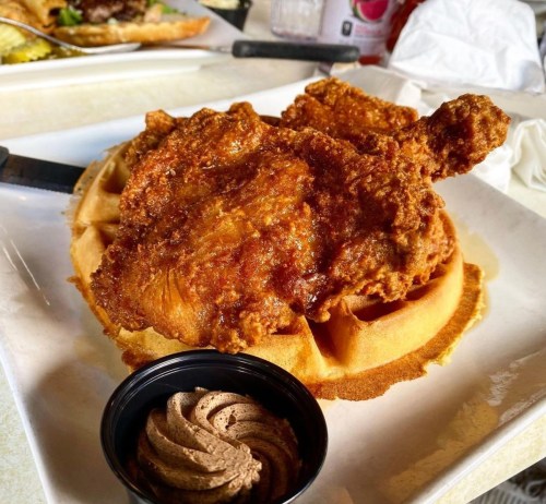 A plate featuring a crispy fried chicken leg on a waffle, accompanied by a small cup of chocolate spread.