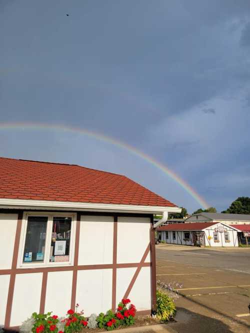 A rainbow arcs over a building with a red roof, framed by a cloudy sky and colorful flowers in the foreground.