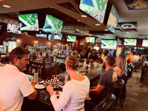 A lively bar scene with patrons watching multiple TVs, enjoying drinks and food at the counter.
