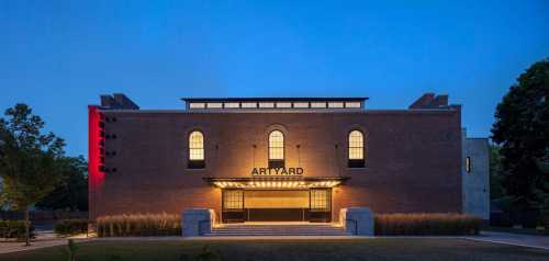 A modern brick building with large windows, illuminated at dusk, featuring the sign "ARTYARD" above the entrance.