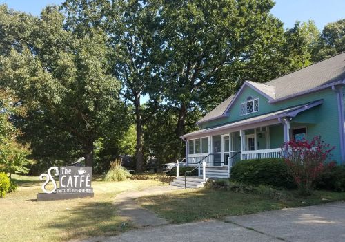 A colorful café building with a sign reading "The Caffè" surrounded by trees and greenery on a sunny day.