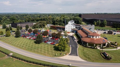 Aerial view of a large building complex with a parking lot filled with cars, surrounded by green fields and trees.