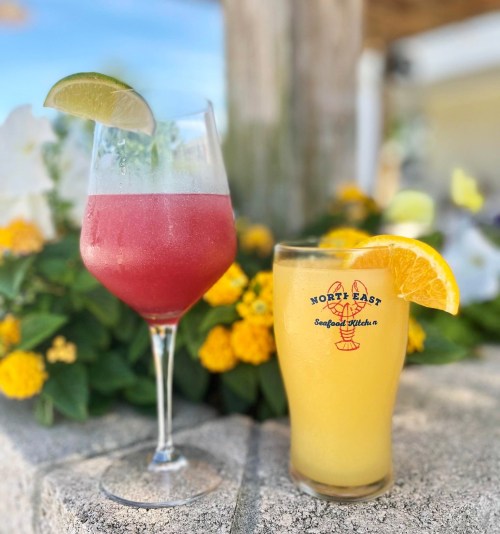 Two colorful cocktails sit on a stone surface, surrounded by vibrant flowers. One is a red drink with a lime, the other is yellow with an orange slice.