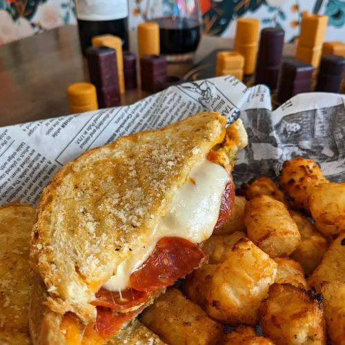 A grilled cheese sandwich with pepperoni, served with crispy tater tots, on a table with colorful game pieces in the background.
