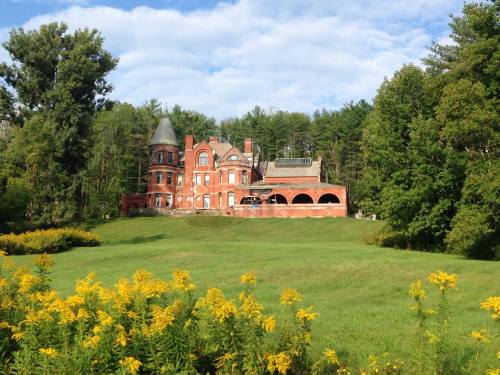 A large red brick mansion surrounded by trees and a grassy field, with yellow flowers in the foreground.