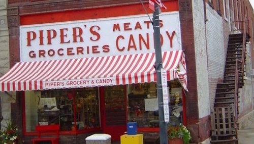 A vintage grocery store with a striped awning, featuring signs for meats, groceries, and candy.