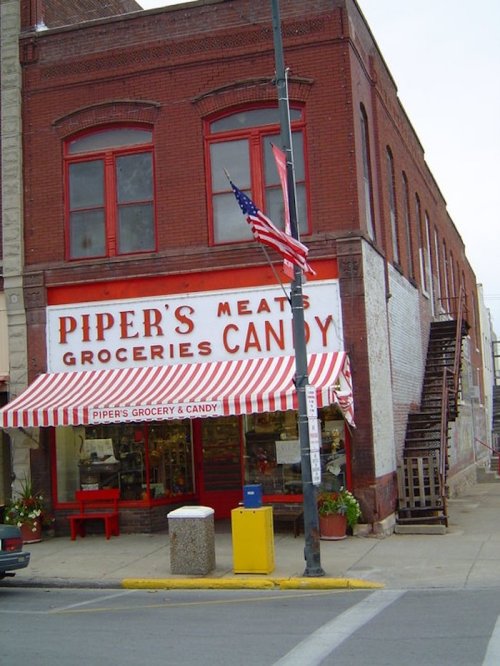 A brick building with a striped awning, displaying "Piper's Meats, Groceries, Candy," and an American flag.