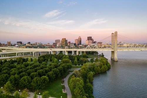 A scenic view of a city skyline at sunset, with a bridge over a river and lush green parks in the foreground.