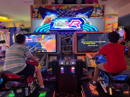 Two children playing a racing arcade game, focused on the screens in front of them. Bright colors and game graphics visible.