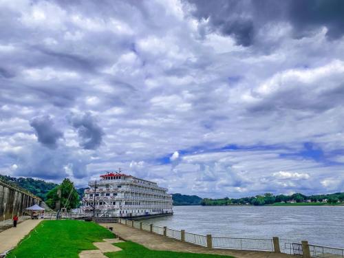 A riverboat docked by the shore under a cloudy sky, with green grass and hills in the background.