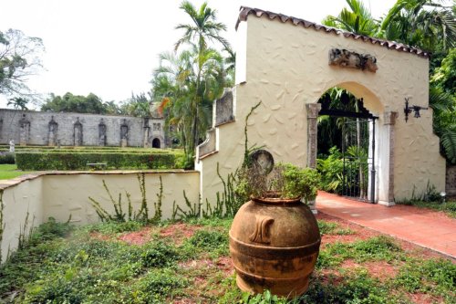 A decorative archway leads to a garden, featuring lush greenery and a large clay pot in the foreground.
