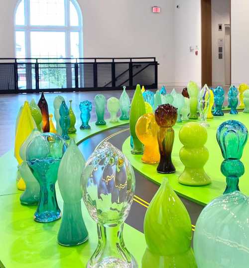 Colorful glass sculptures in various shapes arranged on a bright green circular platform, with large windows in the background.