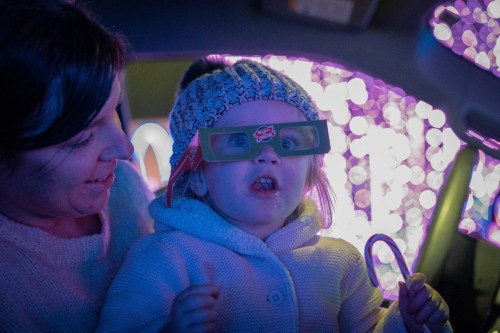 A child wearing 3D glasses sits on a woman's lap, both enjoying colorful holiday lights in the background.