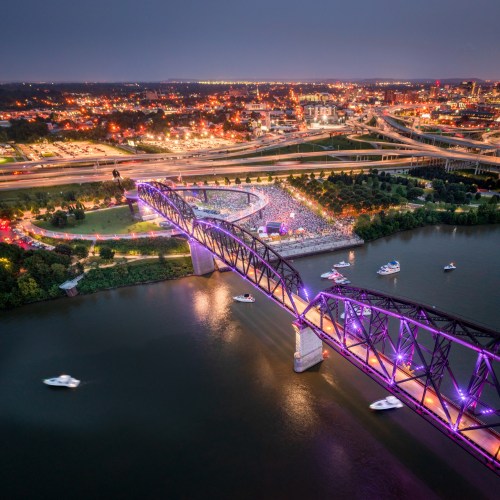 Aerial view of a vibrant cityscape at dusk, featuring a lit bridge, a crowd gathering, and boats on the water.