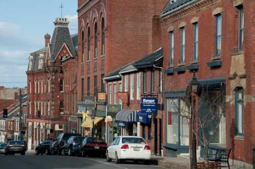 A street view of a historic town with brick buildings, shops, and parked cars under a clear blue sky.