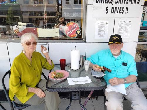 An elderly couple enjoys a meal at an outdoor café, smiling and seated at a small table with plates of food.