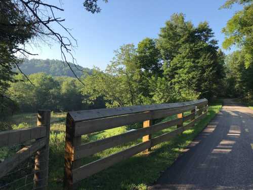 A peaceful rural scene featuring a wooden fence along a winding path, surrounded by lush greenery and distant hills.