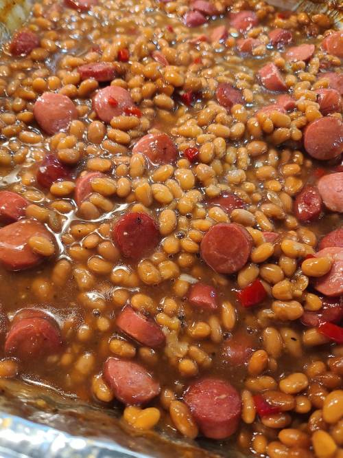 A close-up of baked beans mixed with sliced hot dogs and red peppers in a glossy sauce.