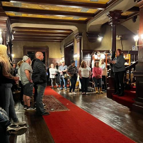 A group of people gathered in a historic room, listening to a speaker on a raised platform. Rich wood decor and red carpet.