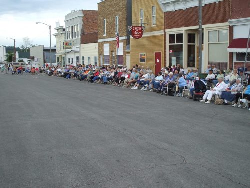 A long line of people sitting in chairs along a street, waiting for an event in a small town.