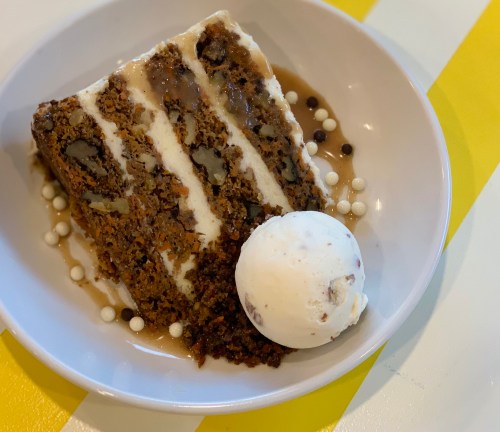 A slice of layered cake with nuts, drizzled with sauce, served with a scoop of vanilla ice cream on a white plate.