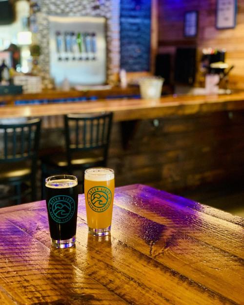 Two glasses of beer, one dark and one light, sit on a wooden table in a cozy bar setting.
