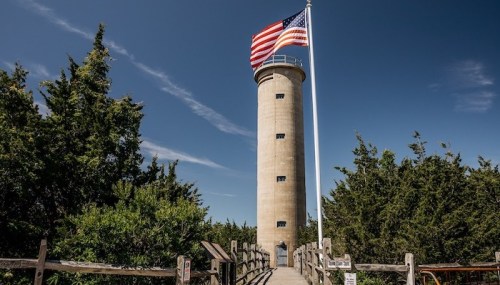 A tall lighthouse with an American flag, surrounded by greenery and a wooden walkway leading to it.