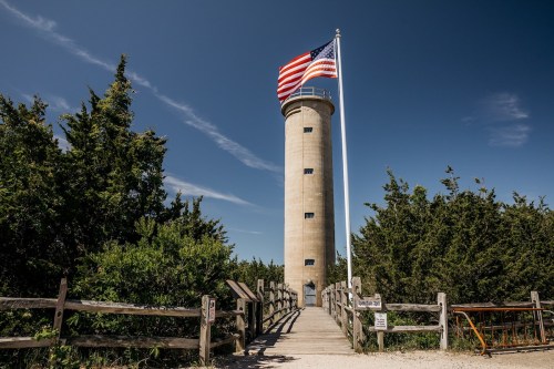 A tall lighthouse with an American flag, surrounded by greenery and a wooden fence under a clear blue sky.