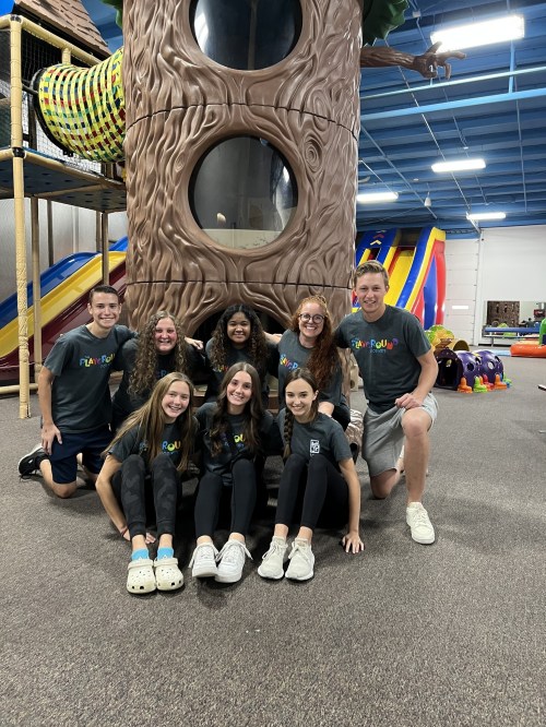 A group of nine people poses in front of a large play structure in a colorful indoor play area.