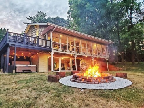 A cozy house with a fire pit in the foreground, surrounded by trees and a grassy area, during sunset.