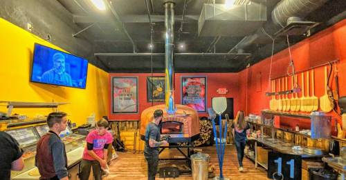 A vibrant pizzeria interior with a wood-fired oven, colorful walls, and staff preparing food. A TV displays a performance.