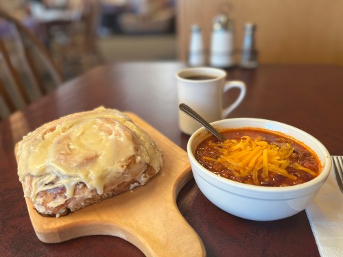 A cinnamon roll with icing on a wooden board next to a bowl of chili topped with cheese and a cup of coffee.