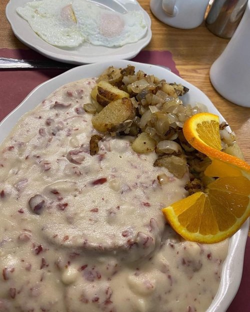 A plate of creamy sausage gravy over biscuits, served with diced potatoes and orange slices, alongside two fried eggs.