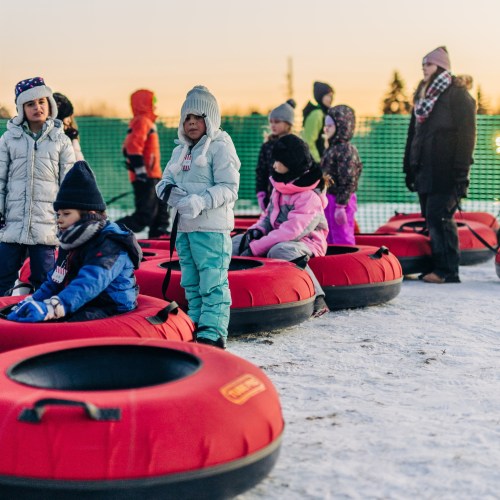 Children in winter clothing sit in red snow tubes on a snowy hill, enjoying a sunny day outdoors.
