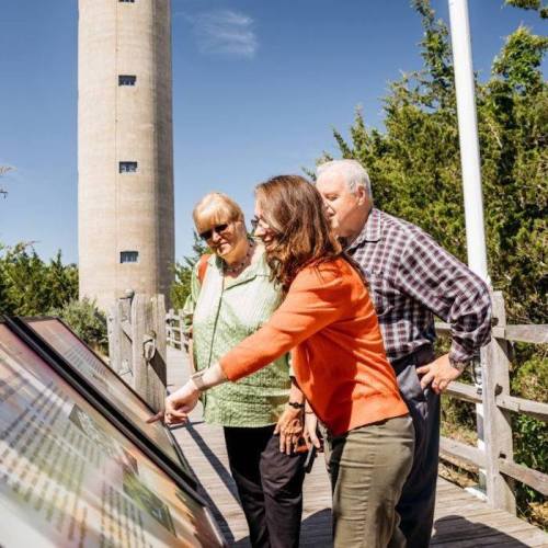 Three people interact with an informational display near a lighthouse, surrounded by greenery on a sunny day.