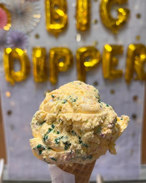 A close-up of a colorful ice cream cone with sprinkles, set against a backdrop of gold "BIG DIPPER" balloons.
