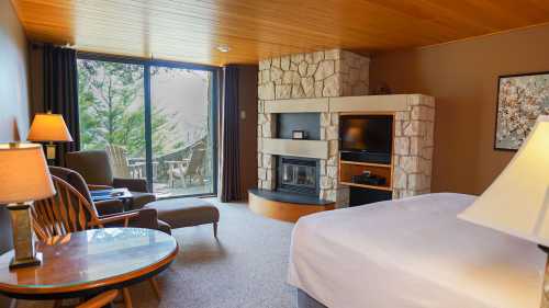 Cozy hotel room with a stone fireplace, seating area, and large windows overlooking a forested view.