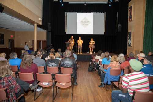 A stage with three speakers presenting to an audience in a theater setting.